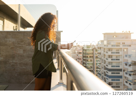Young curly woman standing on balcony overlooking city at sunset with phone in hands, online shopping, social networking, remote work, side view Young curly woman standing on balcony overlooking city at sunset with phone in hands, online shopping, social networking, remote work, side view 127260519