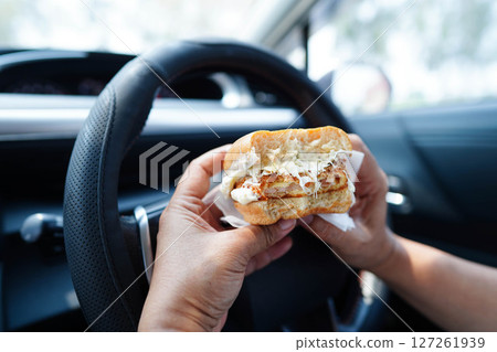 Asian woman driver hold and eat hamburger in car, dangerous and risk an accident. 127261939
