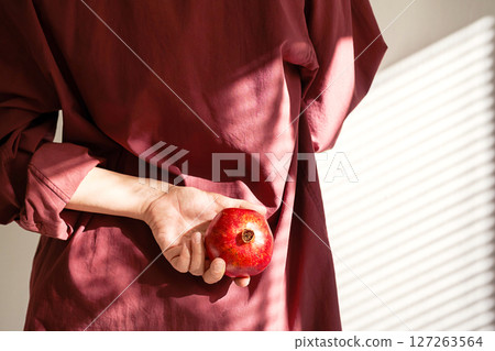 Woman Wearing Burgundy Shirt Holding Red Pomegranate Fruit Behind Back. 127263564