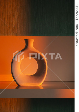 Modern Decorative Ceramic Vase with Circular Hole Stands on Shelf, Illuminated by Warm Orange Light. Home Decor and Style for Interior Design. Modern Decorative Ceramic Vase with Circular Hole Stands on Shelf, Illuminated by Warm Orange Light. Home Decor and Style for Interior Design. 127263610