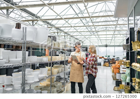 A shop assistant discusses plant pots with a customer in a store. The shelves are filled with diverse, stylish flowerpots. A shop assistant discusses plant pots with a customer in a store. The shelves are filled with diverse, stylish flowerpots. 127265300
