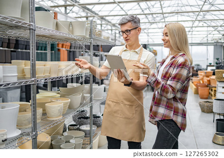 A Caucasian woman discusses flower pots with a shop assistant at a plant store. They're examining options on a tablet device near shelves. 127265302