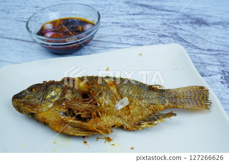 A perfectly golden-fried Mujair or Tilapia rests on a white rectangular plate, with a small bowl of dark dipping sauce in the background. A perfectly golden-fried Mujair or Tilapia rests on a white rectangular plate, with a small bowl of dark dipping sauce in the background. 127266626