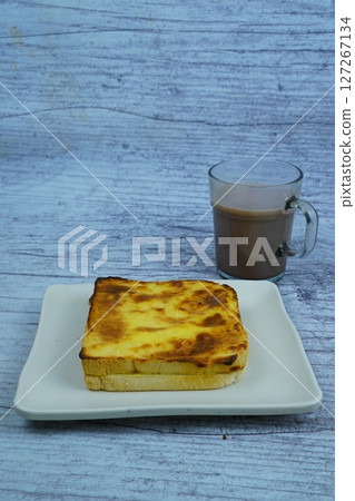 A golden-brown baked toast with a creamy topping is served on a white plate next to a glass of chocolate milk, all on a rustic wooden background. 127267134