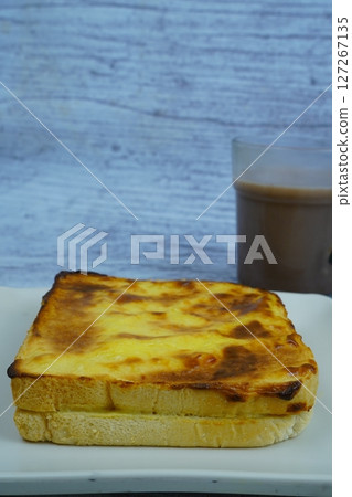 A golden-brown baked toast with a creamy topping is served on a white plate next to a glass of chocolate milk, all on a rustic wooden background. A golden-brown baked toast with a creamy topping is served on a white plate next to a glass of chocolate milk, all on a rustic wooden background. 127267135
