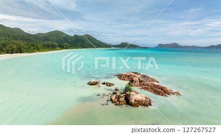 Gentle waves surround rocky outcrops amidst tranquil turquoise waters, with lush hills and a sandy shoreline in the distance. Anse Volbert Beach. Praslin, Seychelles. Gentle waves surround rocky outcrops amidst tranquil turquoise waters, with lush hills and a sandy shoreline in the distance. Anse Volbert Beach. Praslin, Seychelles. 127267527