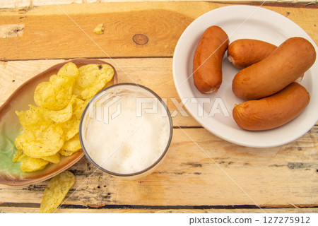 An overhead view of a simple rustic lunch of beer, sausages, potato chips on a light colored wooden background 127275912
