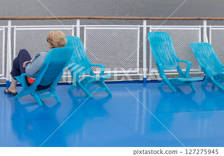 A lone middle-aged woman sits in a chaise lounge on the deck of a cruise ship, looking out to sea. Unrecognizable face 127275945
