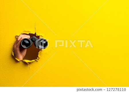 Surveillance concept, hand holding binoculars breaking through yellow wall 127278110
