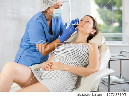 Doctor in disposable mask performs procedure of lip augmentation of girl patient in clinic. Doctor in disposable mask performs procedure of lip augmentation of girl patient in clinic. 127287195