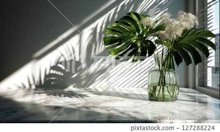 Green Monstera Leaves and White Flowers in Glass Vase on Marble Table 127288224