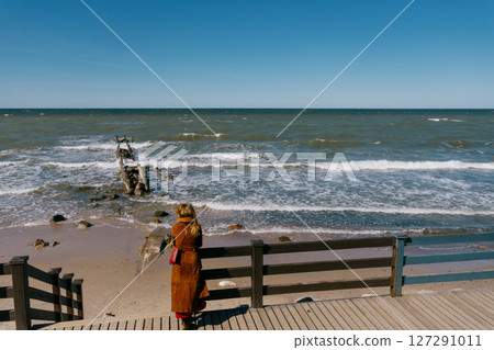 woman in a brown coat stands on a pier overlooking the ocean and enjoys the views.  127291011