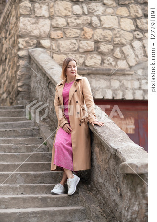 Woman Stairs Fashion: Smiling woman poses on stone stairs with trenchcoat, day, for outdoor photoshoot. 127291091