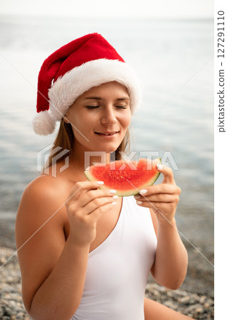 A woman is eating a watermelon while wearing a red santa hat. Scene is lighthearted and festive, as the woman is enjoying a refreshing snack on a sunny day. 127291110