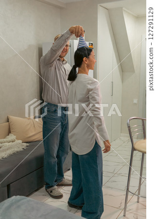 Here's a title following your specifications: Party Hat Man Woman Indoor: Couple playfully placing party hat indoors, likely celebrating birthday, inside home. 127291129