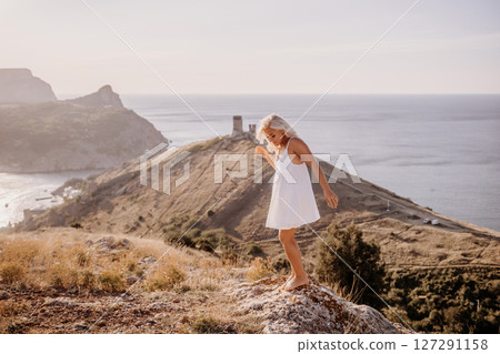 A blonde woman stands on a hill overlooking the ocean. She is wearing a white dress and she is enjoying the view. A blonde woman stands on a hill overlooking the ocean. She is wearing a white dress and she is enjoying the view. 127291158