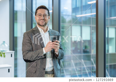Smiling young male businessman in a suit and glasses standing in the office, holding a mobile phone and smiling at the camera Smiling young male businessman in a suit and glasses standing in the office, holding a mobile phone and smiling at the camera 127291669