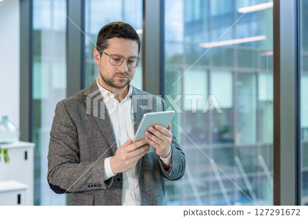 Office worker, young man in a suit standing indoors and using a tablet. Office worker, young man in a suit standing indoors and using a tablet. 127291672
