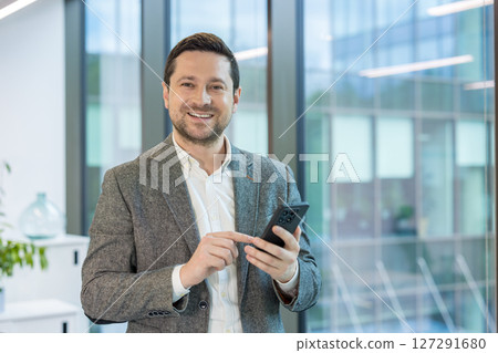 Portrait of a smiling young male businessman standing in the office in a business suit, holding a mobile phone and looking at the camera. 127291680