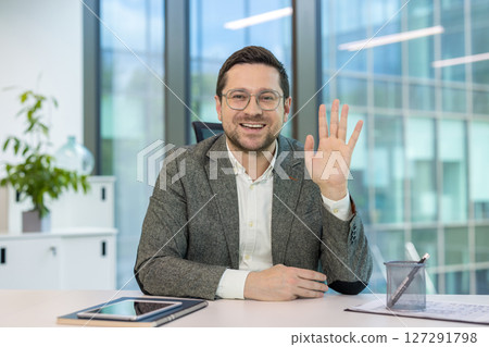 Smiling young man sitting at his workplace in the office, greeting and waving at the camera. 127291798