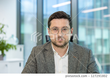 Portrait of a young male businessman sitting in the office at his workplace and looking seriously at the camera. Portrait of a young male businessman sitting in the office at his workplace and looking seriously at the camera. 127291802