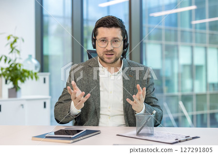 Portrait of a young man in a headset and computer sitting in an office at a table and holding an online video conference, explaining by gesturing with his hands. 127291805