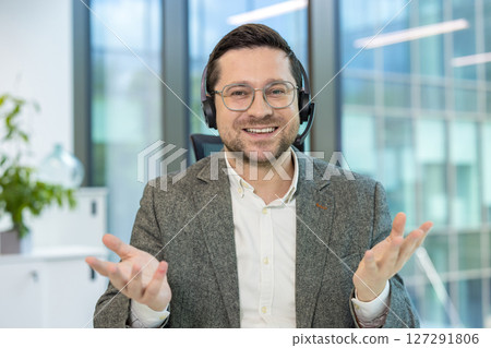 Portrait of a smiling young man in a headset sitting at an office desk and talking to the camera, while gesturing with his hands. Portrait of a smiling young man in a headset sitting at an office desk and talking to the camera, while gesturing with his hands. 127291806