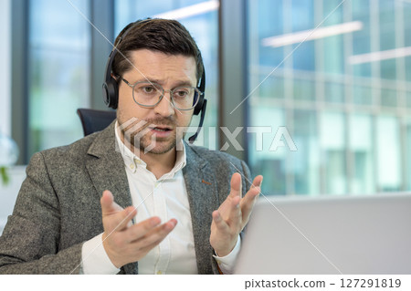 A man wearing a headset speaks in front of a laptop, appearing engaged in a virtual meeting at the office. 127291819