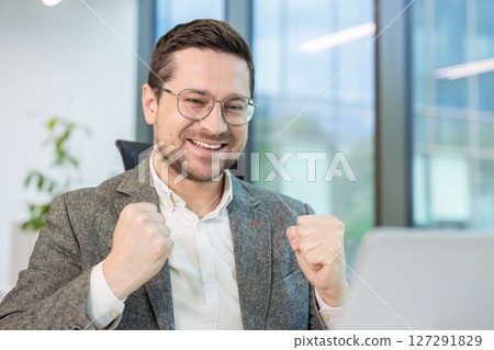 A successful businessman celebrates with fists clenched, radiating joy and triumph. He wears glasses and a blazer, appearing happy in his office. A successful businessman celebrates with fists clenched, radiating joy and triumph. He wears glasses and a blazer, appearing happy in his office. 127291829