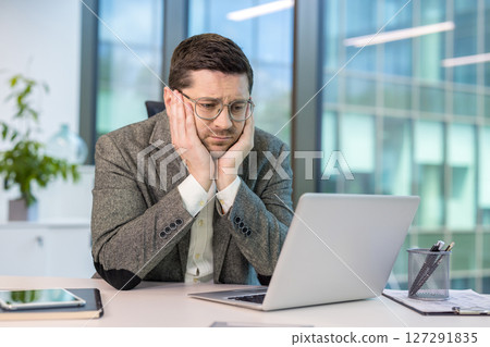 A tired and depressed young male office worker in a suit sits at his desk and looks boredly at the laptop screen. A tired and depressed young male office worker in a suit sits at his desk and looks boredly at the laptop screen. 127291835