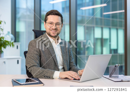 A smiling businessman working on a laptop in a modern office setting, looking directly at the camera. 127291841