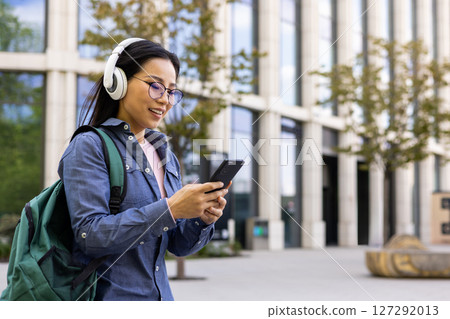 An asian woman walks and looks at her phone, wearing headphones and a backpack. She smiles while checking her smartphone in front of a modern building. An asian woman walks and looks at her phone, wearing headphones and a backpack. She smiles while checking her smartphone in front of a modern building. 127292013