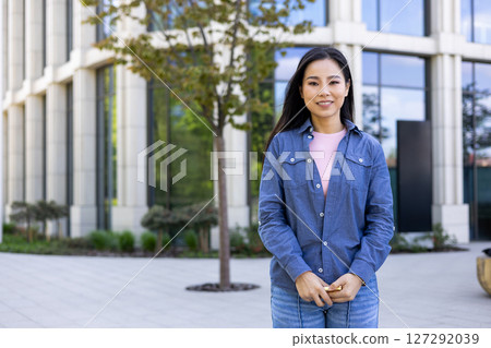 A smiling asian woman poses outside a modern building. She wears a denim shirt and jeans, looking at the camera with confidence. 127292039