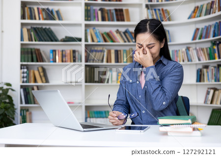 An asian woman rubs her tired eyes while studying at a library. She sits at a desk with a laptop, glasses, and books. An asian woman rubs her tired eyes while studying at a library. She sits at a desk with a laptop, glasses, and books. 127292121