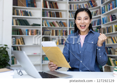An excited asian woman holds a letter, celebrating success in a library setting, with books in the background. A laptop is also on the desk. 127292129