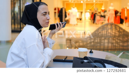 Young muslim woman wearing traditional hijab sitting at table in a shopping mall food court, talking on mobile phone and drinking coffee, enjoying a break from shopping. Free from prejudice of Muslim 127292681