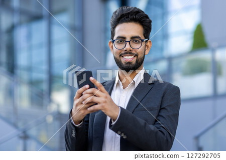 Close-up portrait of a young Indian male businessman standing near office buildings, holding a phone and smiling at the camera. Close-up portrait of a young Indian male businessman standing near office buildings, holding a phone and smiling at the camera. 127292705