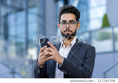 Close-up photo of a young Indian businessman man standing outside in a suit and glasses, holding a phone and looking seriously at the camera. Close-up photo of a young Indian businessman man standing outside in a suit and glasses, holding a phone and looking seriously at the camera. 127292706