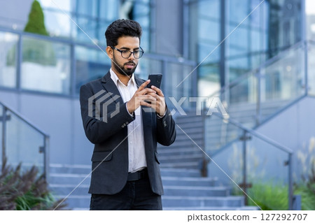 A young Indian businessman in a suit is standing outside an office building and using a mobile phone, typing, reading a message. 127292707