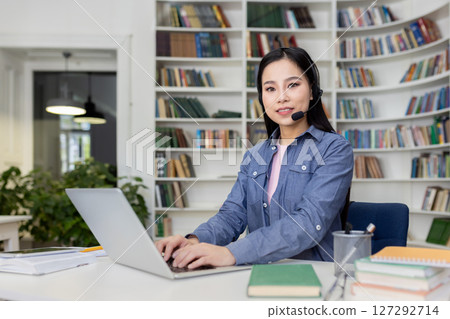 A smiling Asian woman with a headset is working on her laptop in a library setting, focused and attentive. A smiling Asian woman with a headset is working on her laptop in a library setting, focused and attentive. 127292714