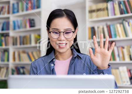 A young woman with glasses smiles while video calling in a library, waving her hand. The backdrop features a bookshelf. 127292738