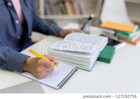 A person studies and takes notes in a book-filled setting, emphasizing education and learning environment. 127292756