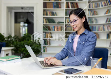 A smiling Asian woman works on a laptop in a library, surrounded by bookshelves, showcasing education and technology use. A smiling Asian woman works on a laptop in a library, surrounded by bookshelves, showcasing education and technology use. 127292757