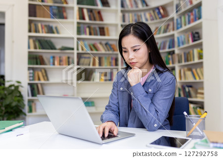 An asian woman works on a laptop in a library, deep in thought with her hand on her chin. Shelves filled with books are in the background. 127292758