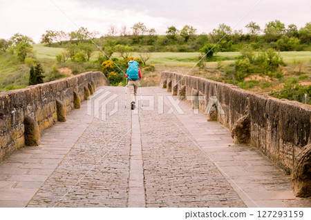 A traveler walks alone across a historic stone bridge in Obanos, Spain, surrounded by nature A traveler walks alone across a historic stone bridge in Obanos, Spain, surrounded by nature 127293159
