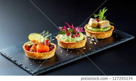 A trio of elegant mini tartlets avocado lime mousse, smoked salmon with pomegranate, and kimchi with tofu beautifully plated on a matte slate board with vibrant colors and gourmet textures. 127293234