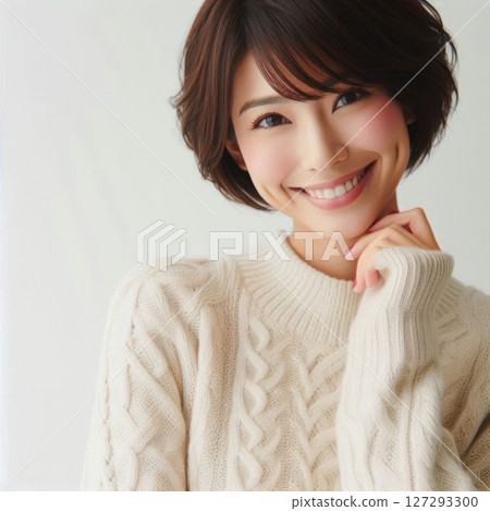 A beautiful Japanese woman with short hair smiles warmly while touching her chin, wearing an elegant sweater and illuminated by soft natural side light. 127293300