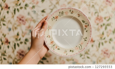 Hand holding vintage porcelain plate with floral design near patterned wallpaper, emotional close-up with historic feel 127293553