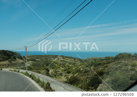 Scenic coastal road with view of Cabo da Roca lighthouse in Sintra Portugal curving highway through natural cliffs overlooking Atlantic Ocean under clear blue sky 127296508
