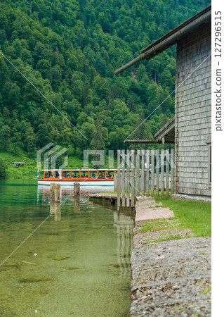 A boat gracefully moves through the calm waters of a beautiful lake, bordered by dramatic green hills and dense forests under a cloudy sky. The peaceful setting invites relaxation. Konigssee Bavaria 127296515